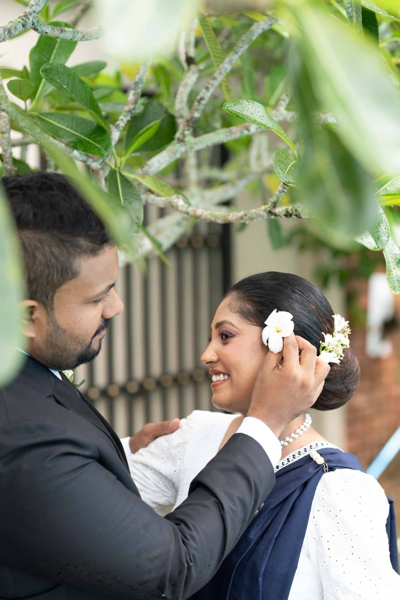 Tender face-to-face moment as the groom tucks a white frangipani flower behind the smiling bride's ear, framed by leaves