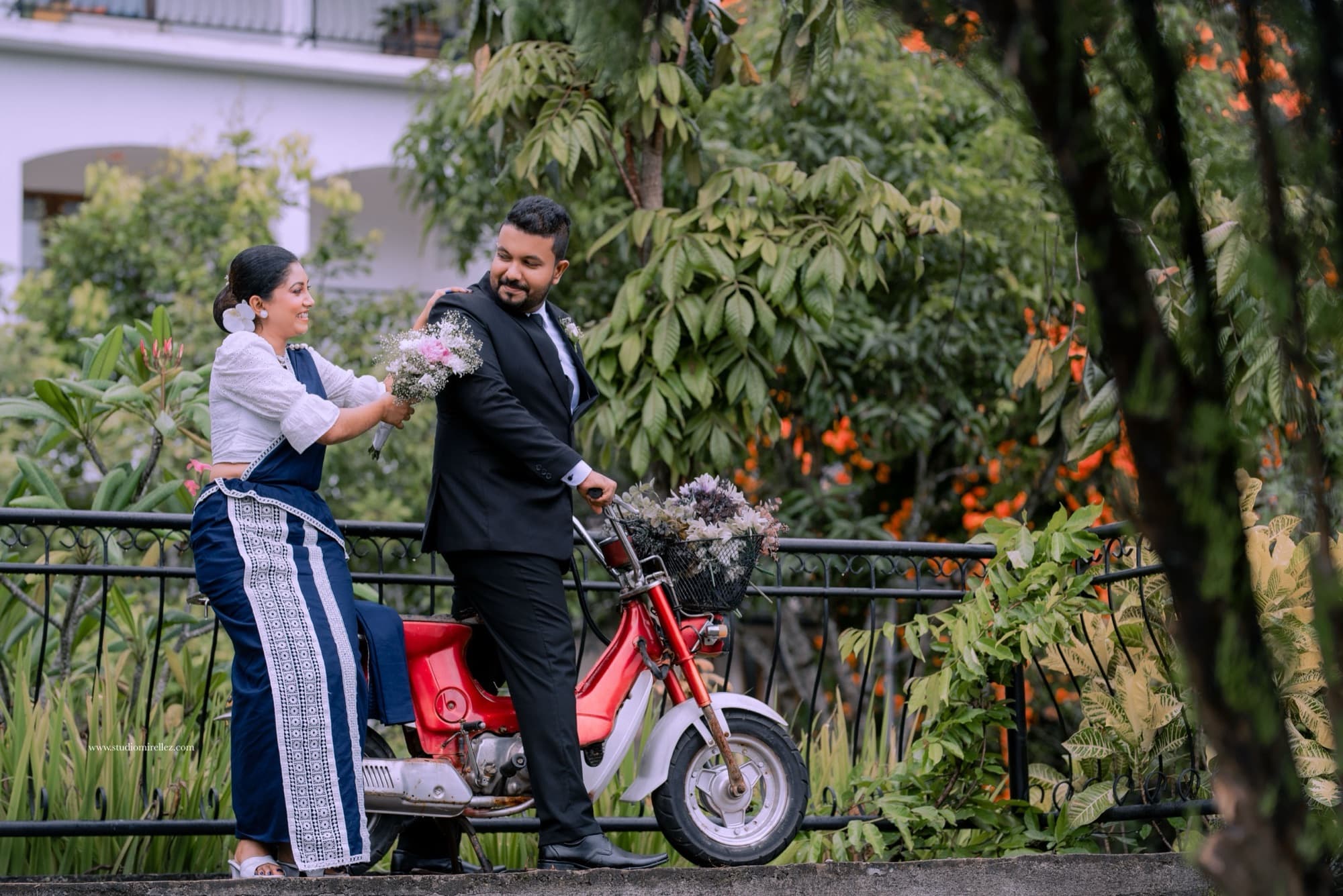 Playful candid of the couple beside a red vintage scooter on a garden bridge, the bride handing over a bouquet amid tropical foliage