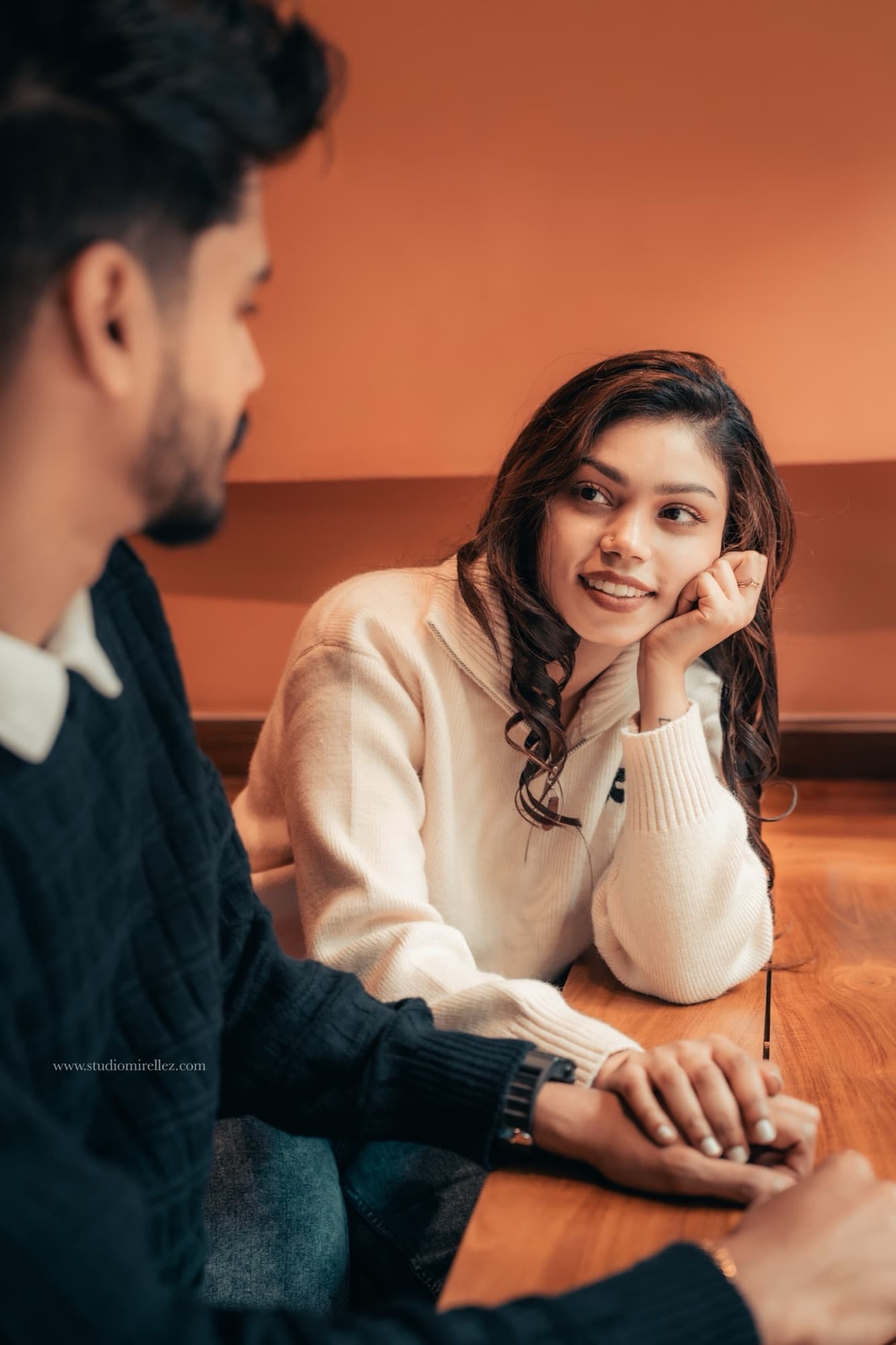 The couple lean across a wooden cafe table holding hands, Mashi resting her chin on her palm and smiling