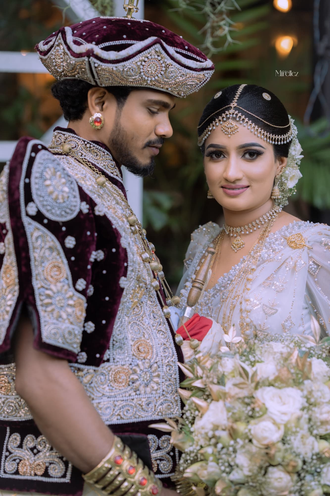 Close portrait of the bride in a beaded white Kandyan saree and pearl headpiece, the groom in a maroon velvet nilame jacket turned toward her