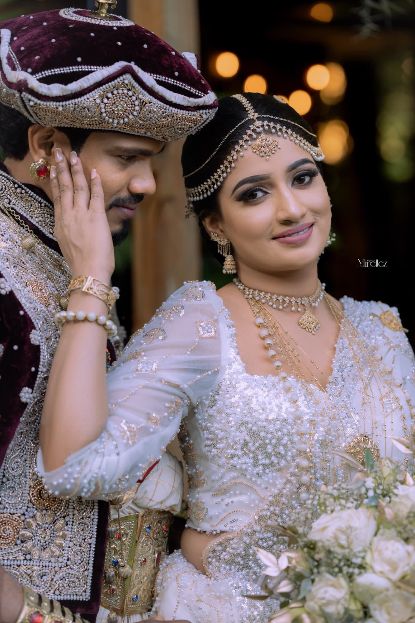 Bride smiling warmly as the groom gently rests his hand against her cheek, gold jewelry catching the light against bokeh