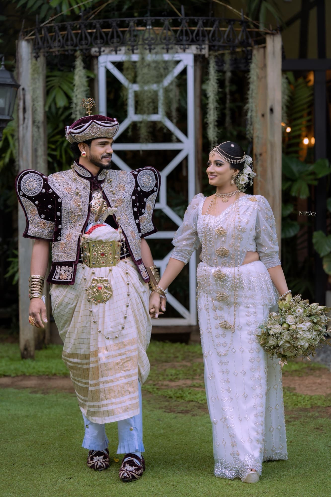 Wide editorial portrait of the couple walking hand in hand across a green lawn beneath a draped garden pavilion