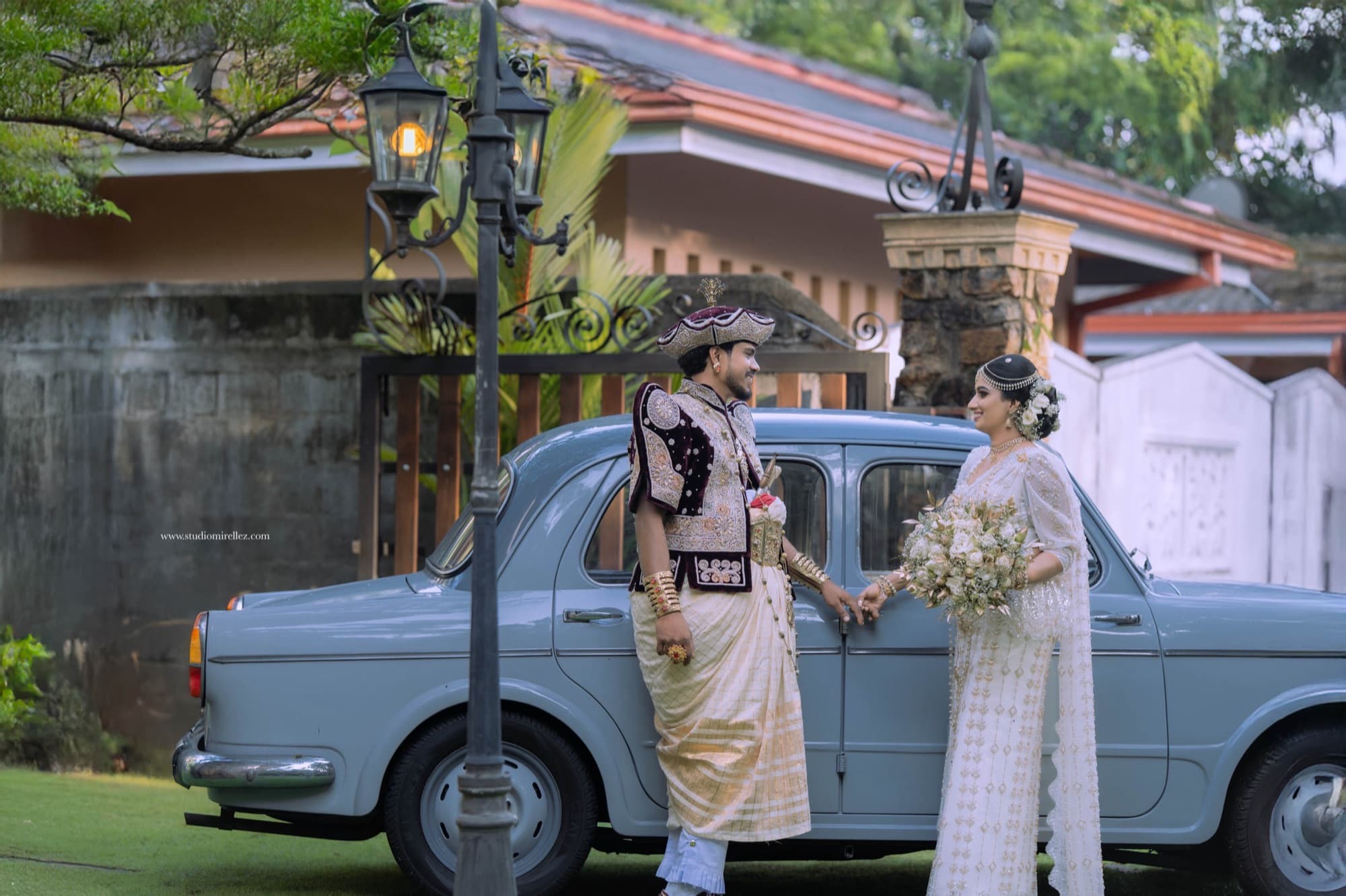 Bride and groom posed beside a vintage pale-blue car in front of a rustic stone house, holding a white bouquet