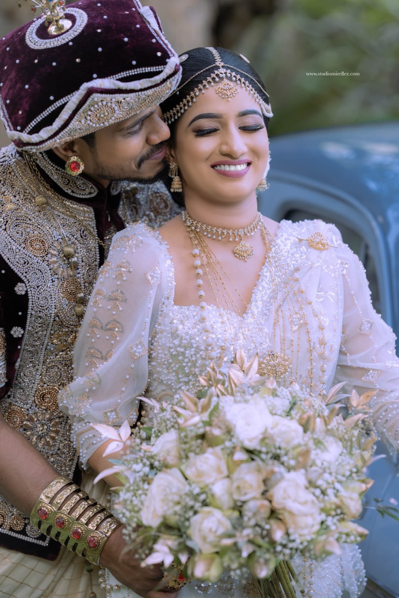 Groom kissing the bride's temple as she smiles with eyes closed, holding a lush white rose and baby's-breath bouquet