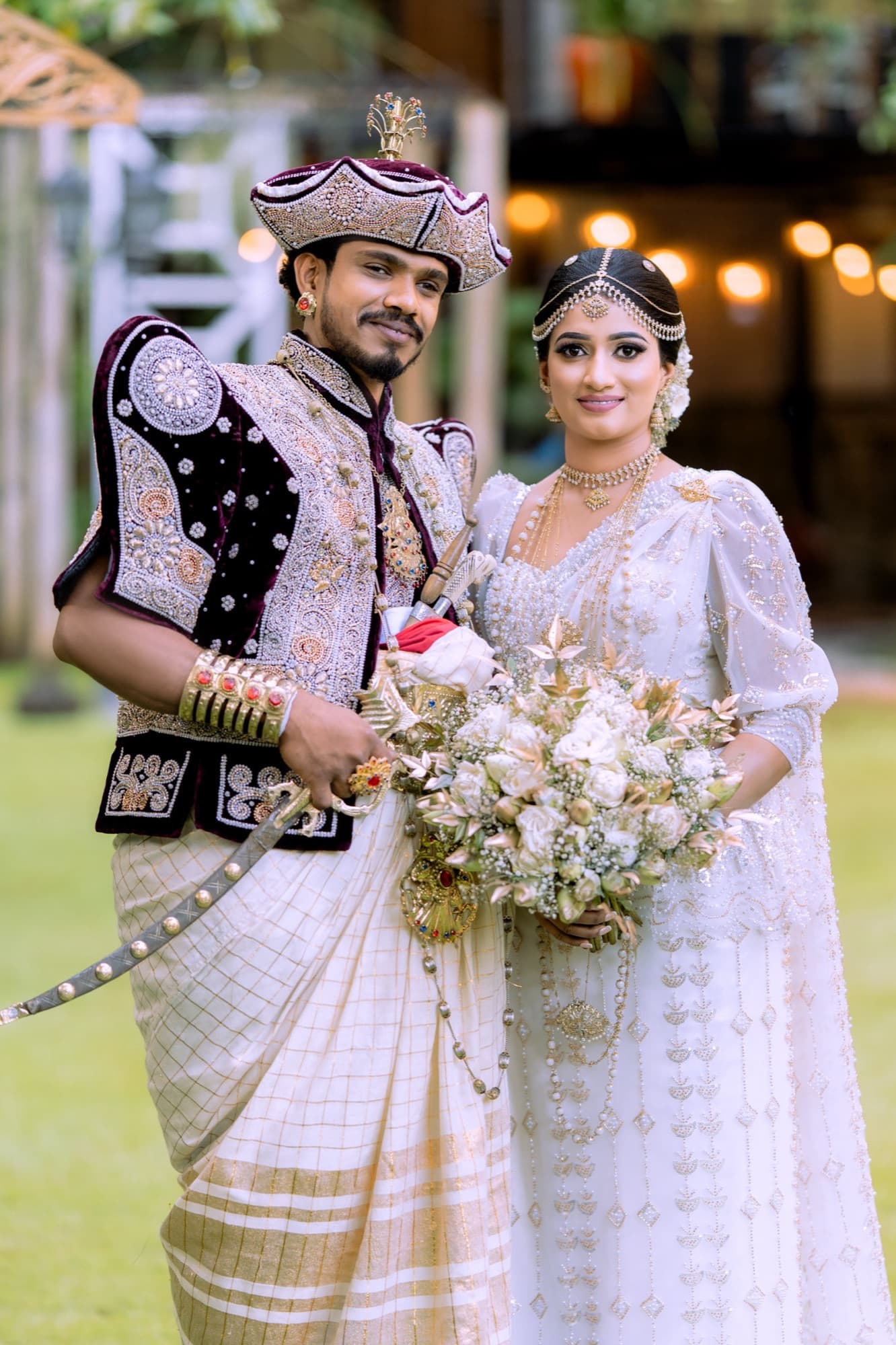 Full-length portrait of the wedding couple in ornate gold-trimmed Kandyan dress against a softly lit garden backdrop