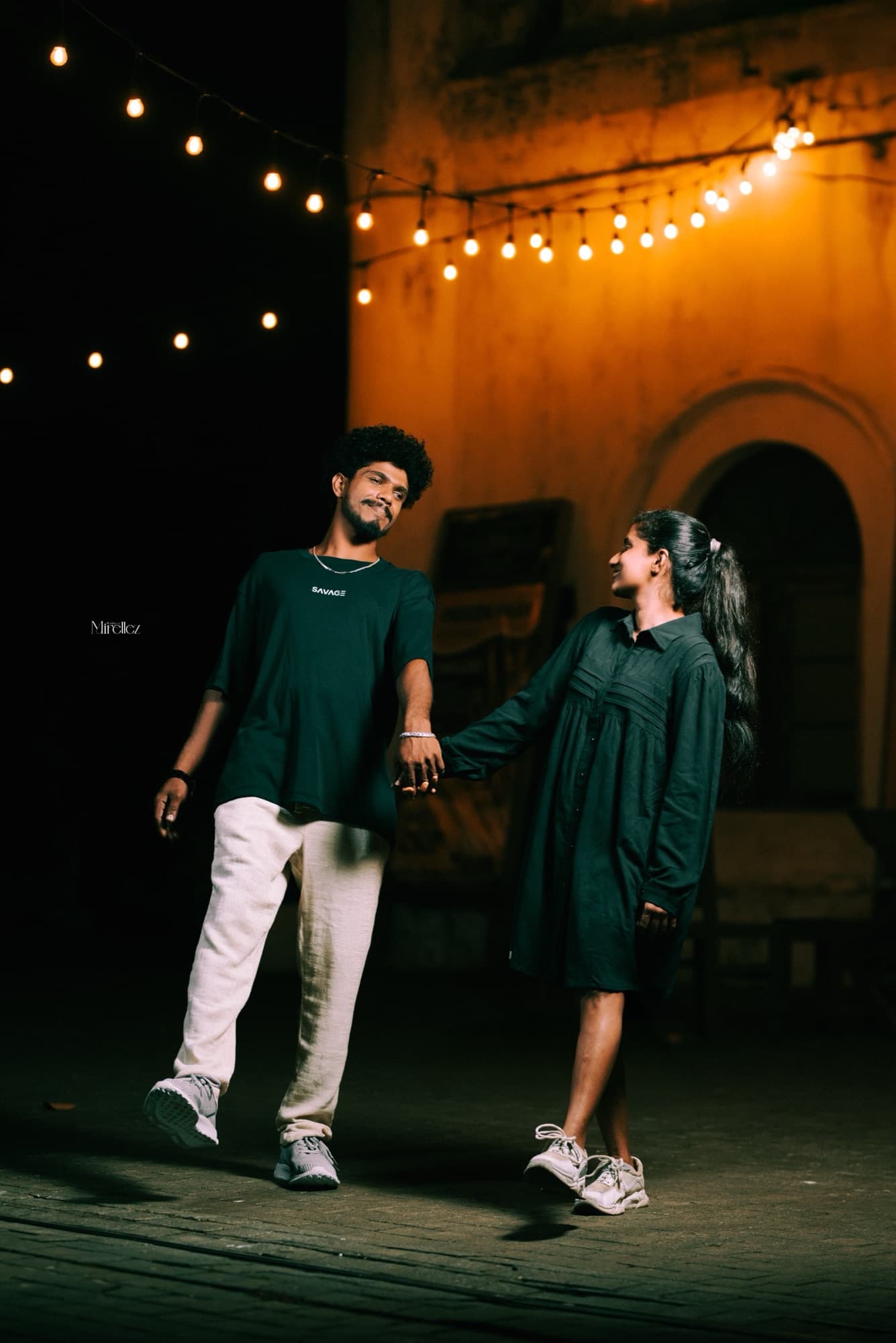 Full-length night shot of the couple holding hands and smiling at each other under glowing string lights against a warm-lit colonial wall.