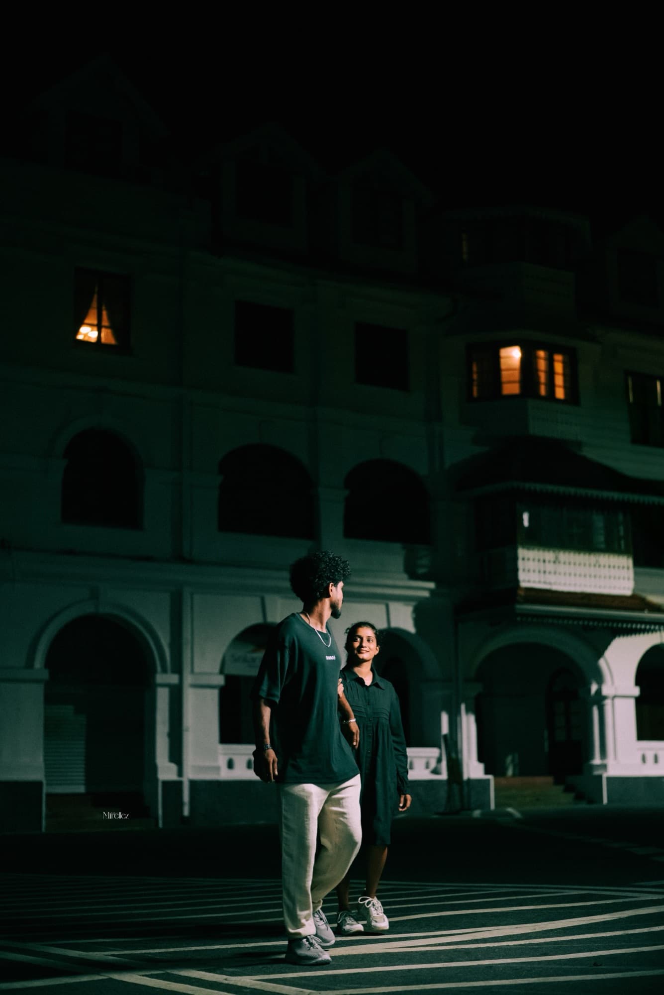 Wide environmental portrait of the couple walking together at night before the illuminated arches of a grand white colonial building.