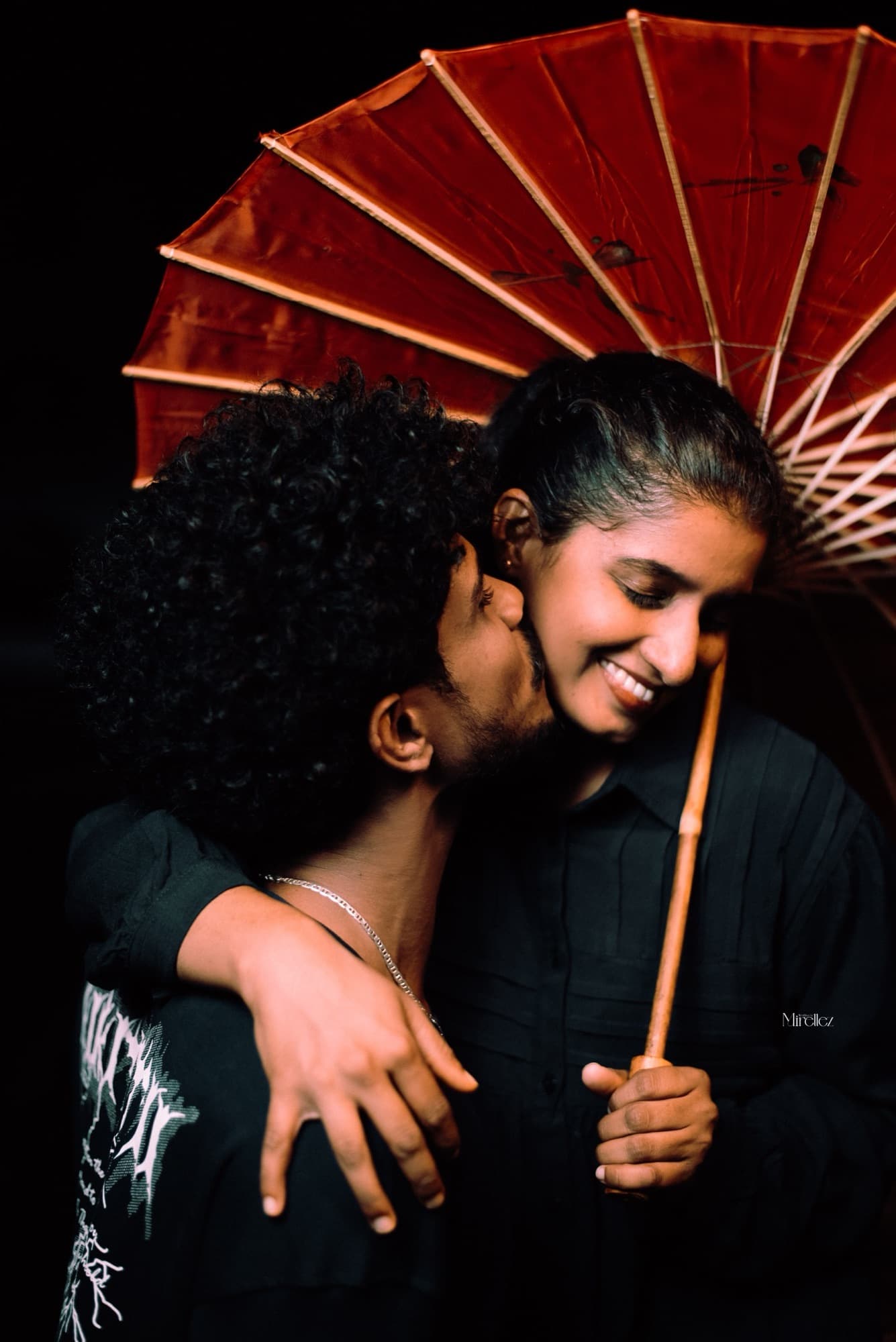 Intimate close-up of the man kissing the smiling woman's cheek beneath a glowing red paper parasol against a black backdrop.