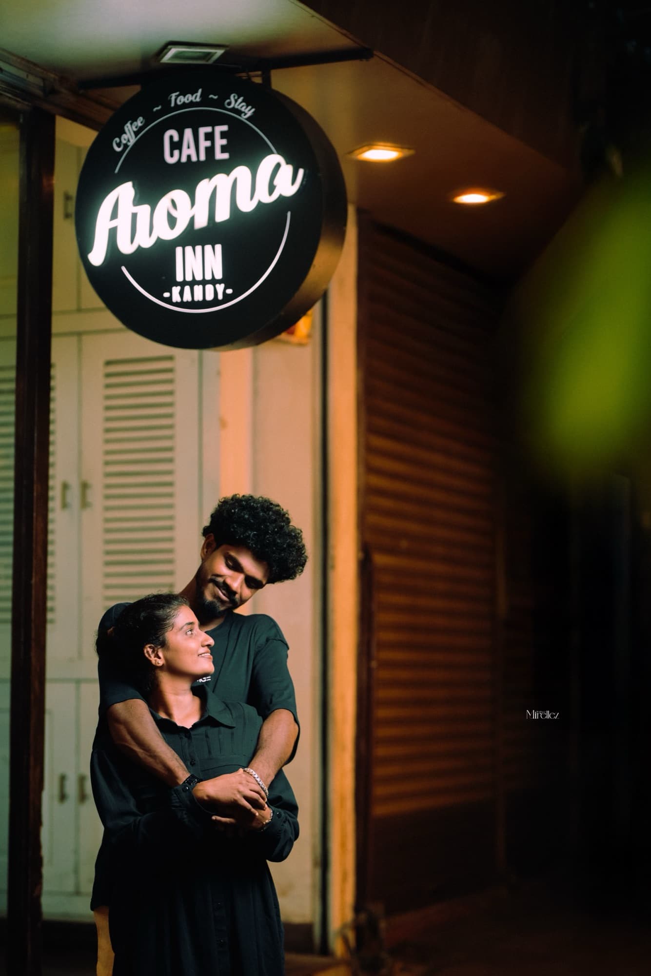 The couple embracing on a Kandy street at night beneath the round illuminated Cafe Aroma Inn sign.