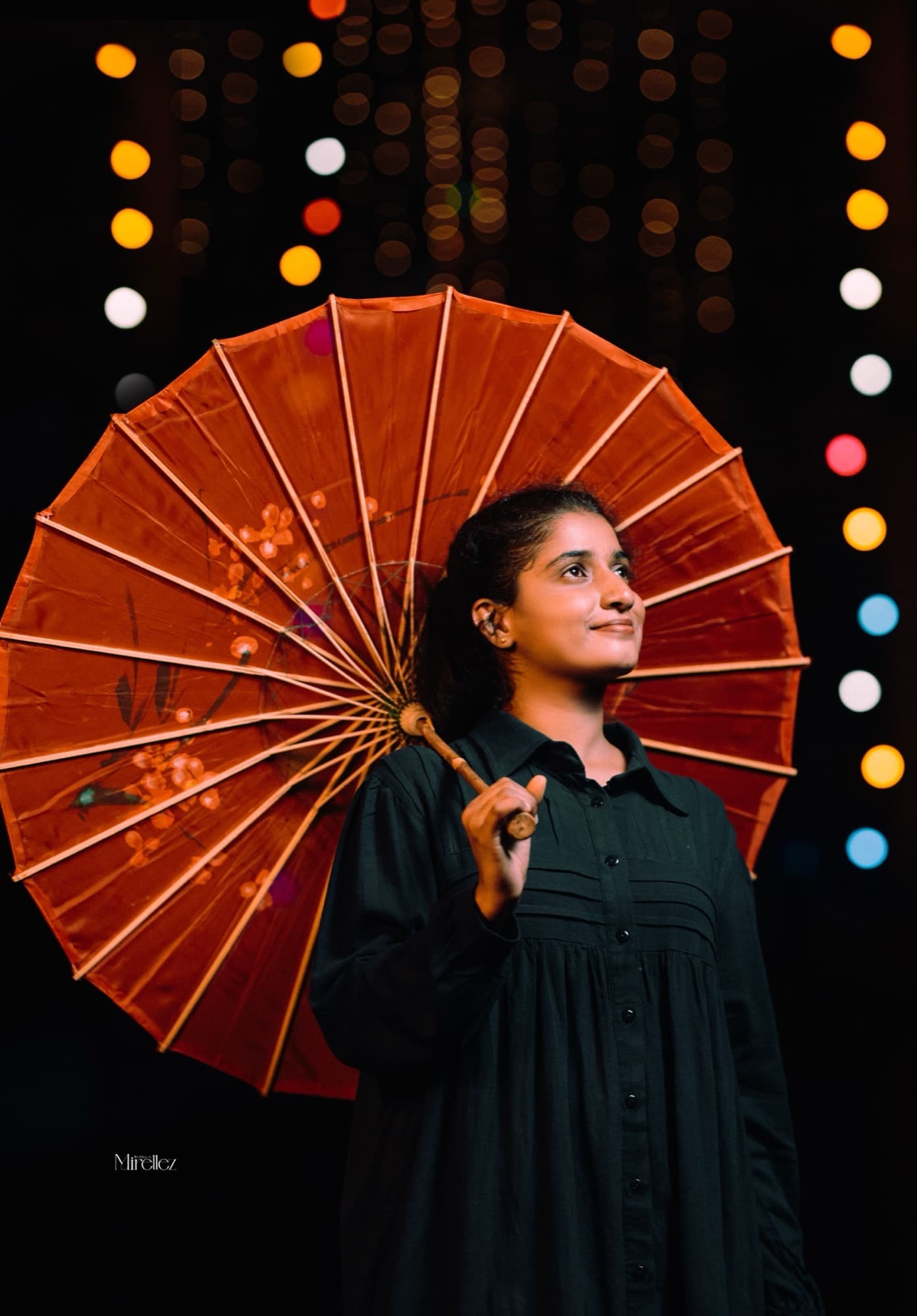 Solo portrait of the woman in a black dress holding an open red parasol, gazing upward amid colourful bokeh lights.
