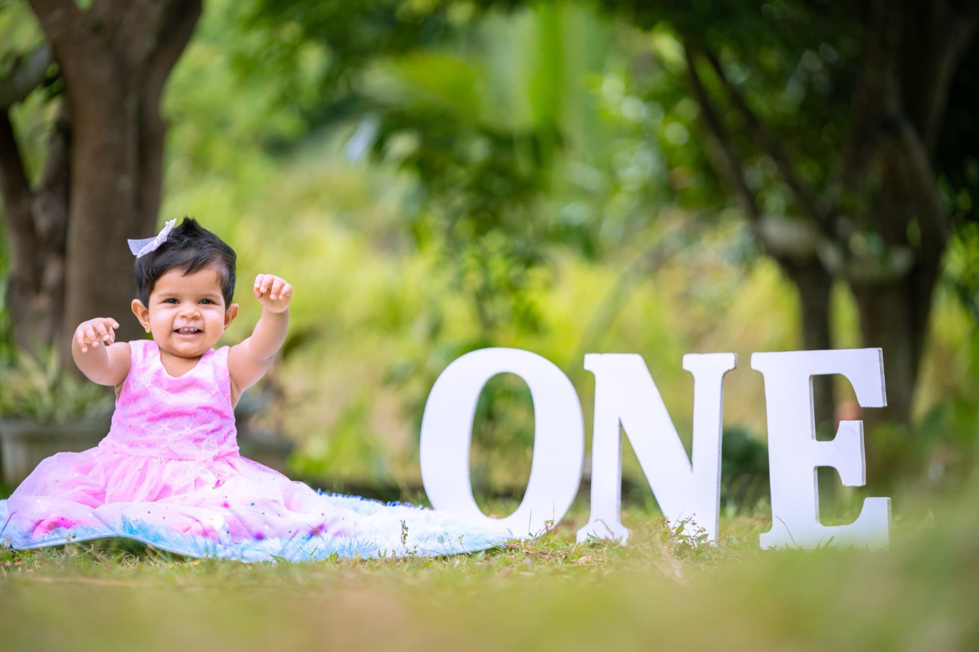 Baby girl in a layered pink tutu dress sits on the grass with arms raised in joy beside oversized white ONE letters in a leafy park.