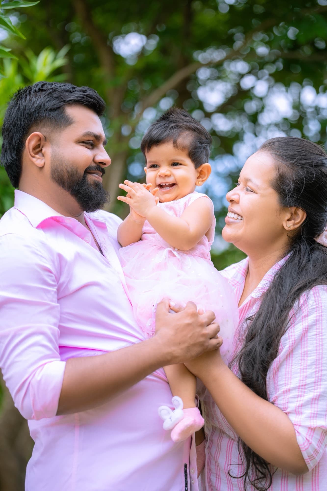 Mother and father hold their laughing baby between them, the family sharing a tender candid moment under soft dappled trees.