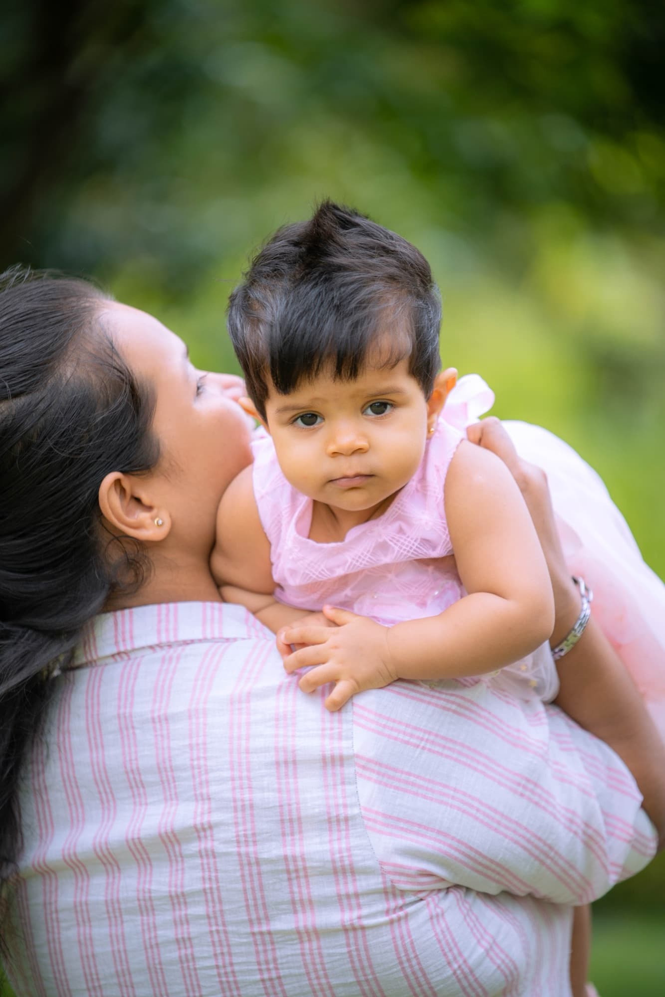 Close portrait of a baby in a pale pink dress resting on her mother's shoulder, gazing calmly at the camera with green bokeh behind.