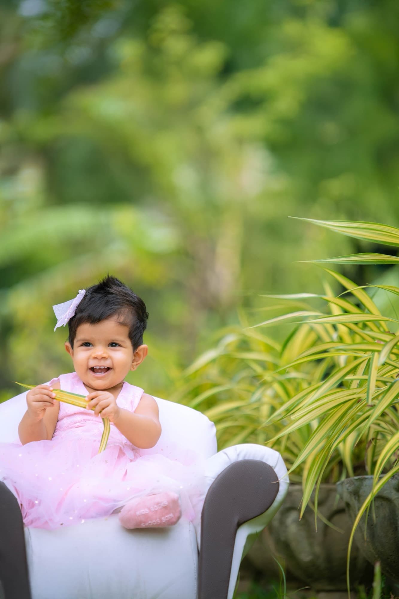Baby in a pink tutu sits in a small white chair holding a leaf, framed by tropical foliage and lush green garden bokeh.