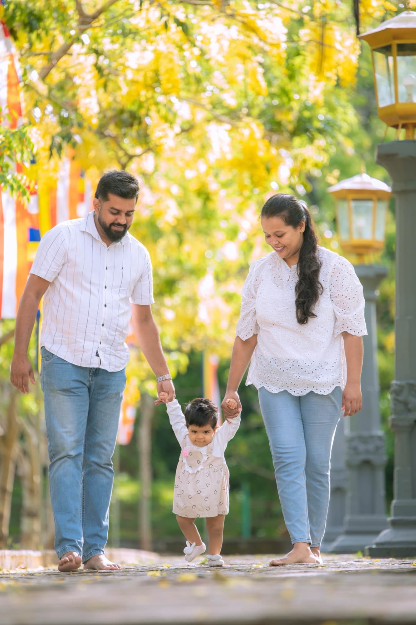 Mother and father in white and denim walk their baby forward by both hands along a temple path strewn with yellow petals.