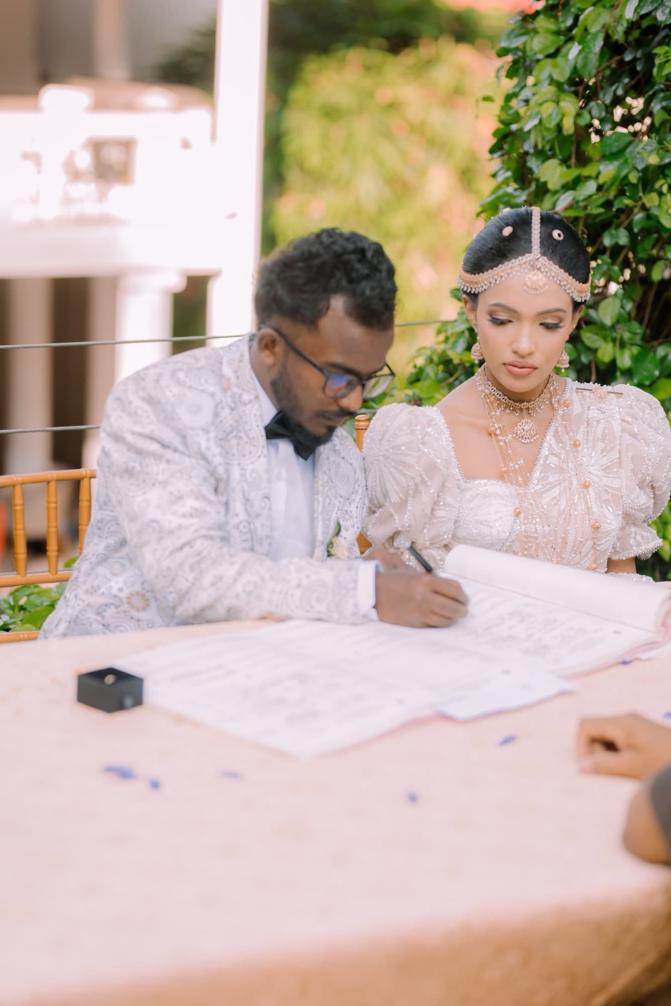 Close intimate moment as the groom signs the marriage register beside the bride in a beaded headpiece, ring box on the soft pink table.