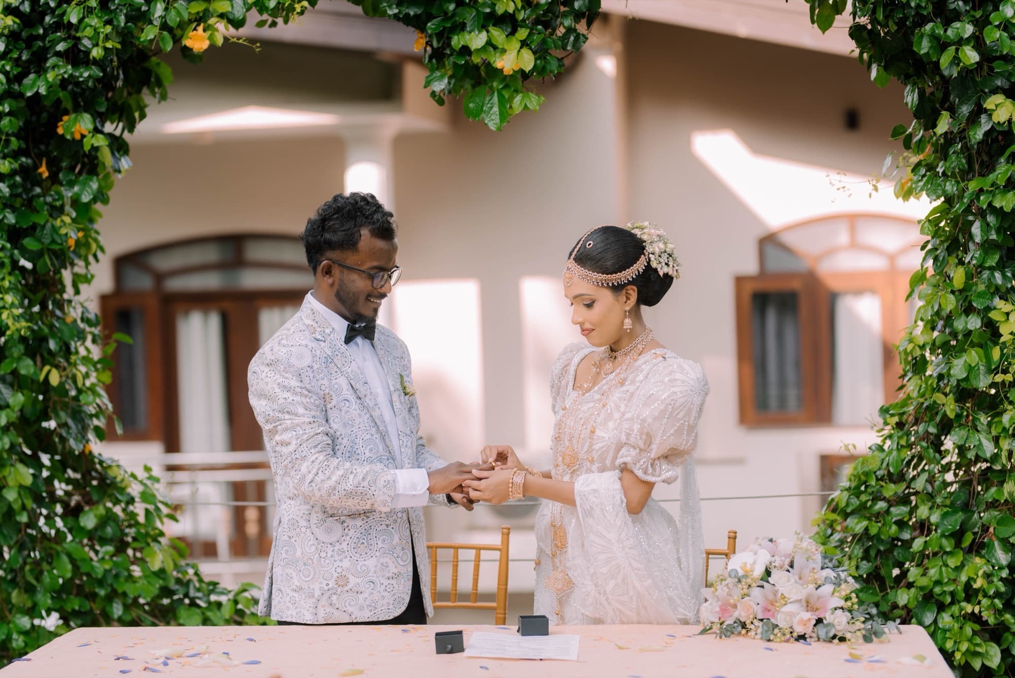 Wide frame of the couple clasping hands during the ring exchange, framed by a green vine arch in front of a colonial villa.