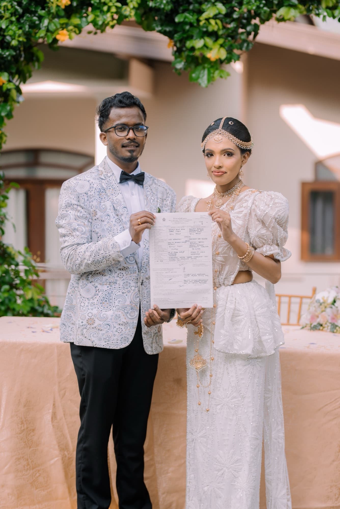 Bride and groom standing together holding up their signed marriage certificate, looking to camera in front of greenery.