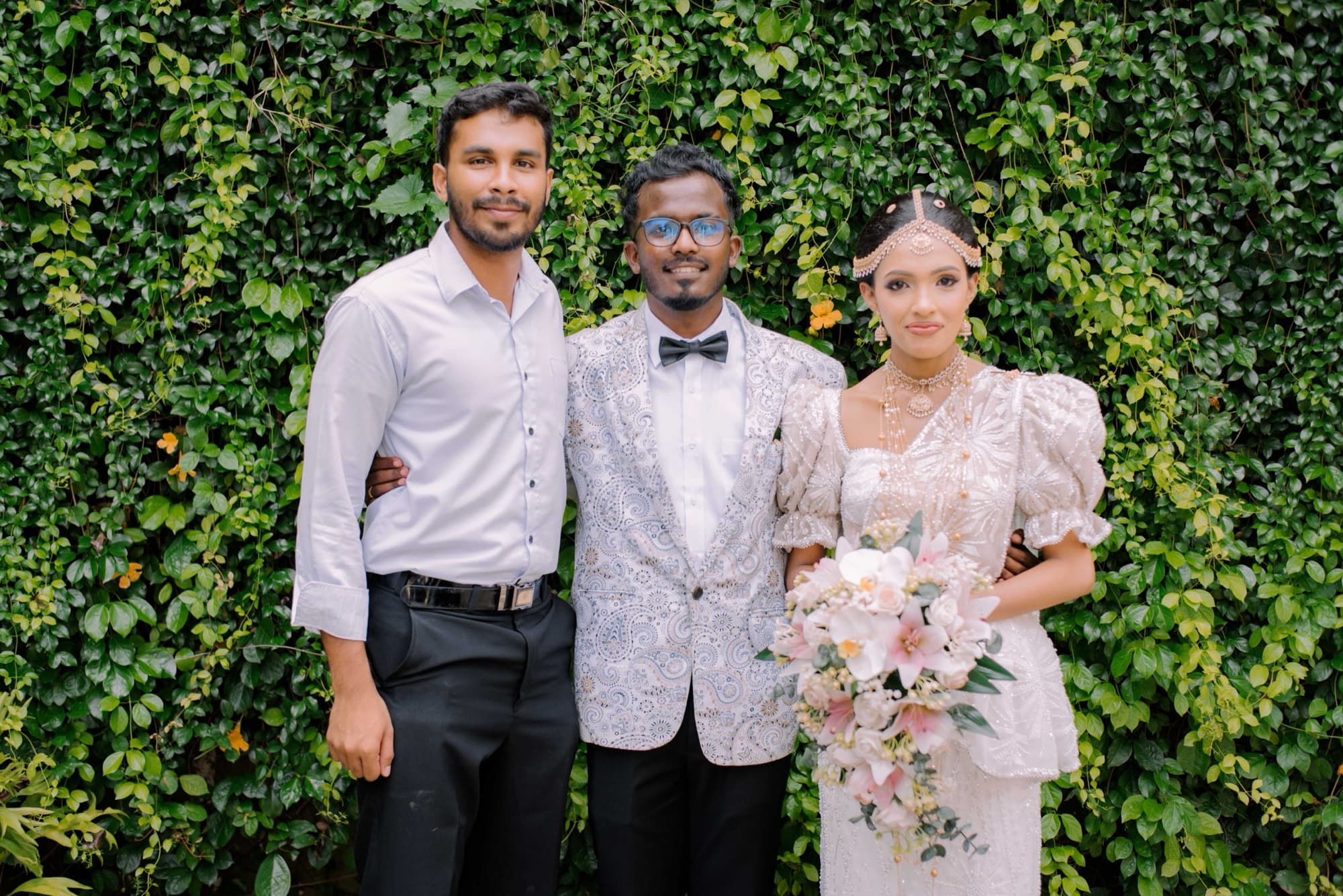 The couple with a friend posed against a lush ivy wall, the bride holding a pastel pink-and-cream bouquet.