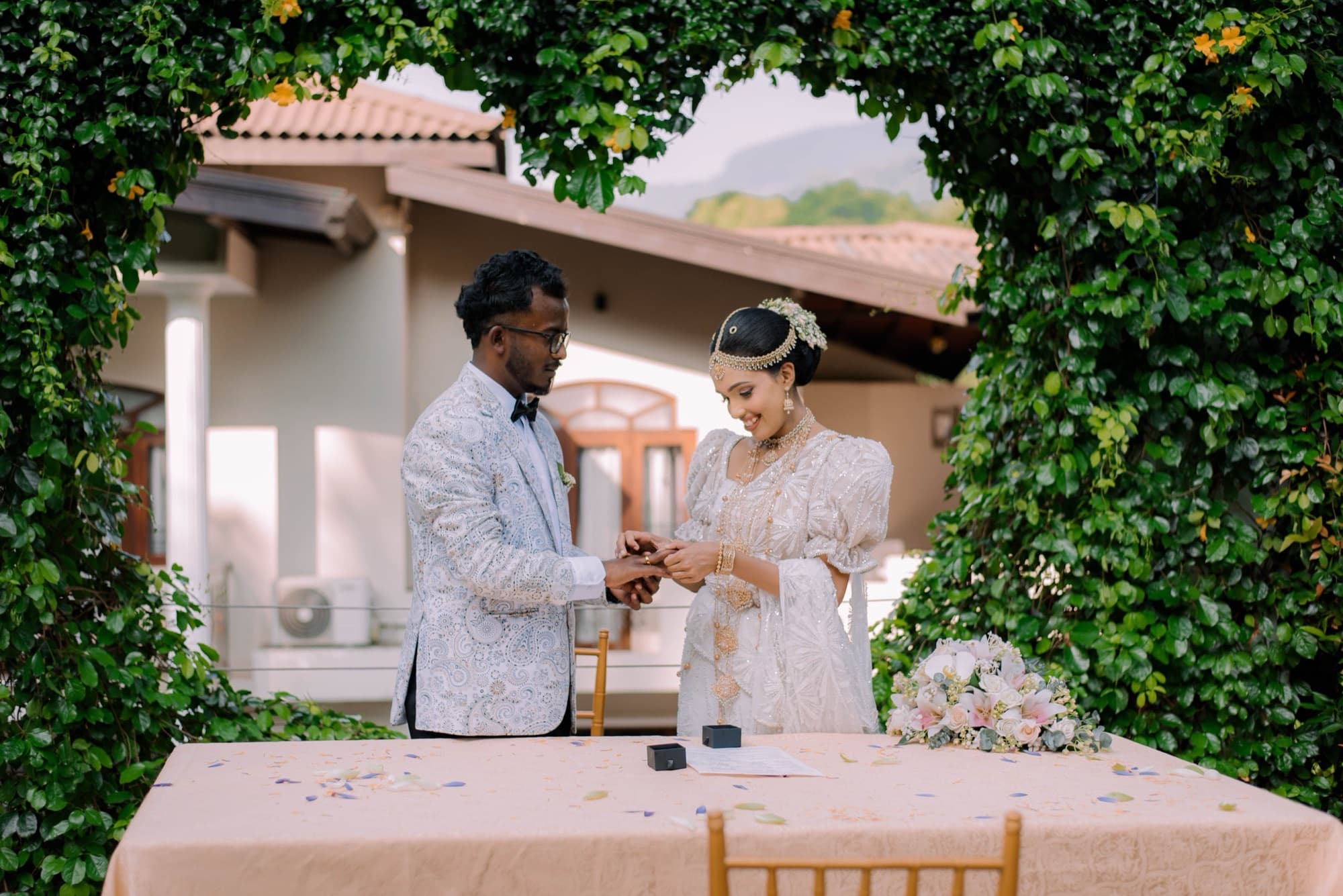 Full-length portrait of the newlyweds raising their hands to show their rings while holding the certificate, mountains beyond.