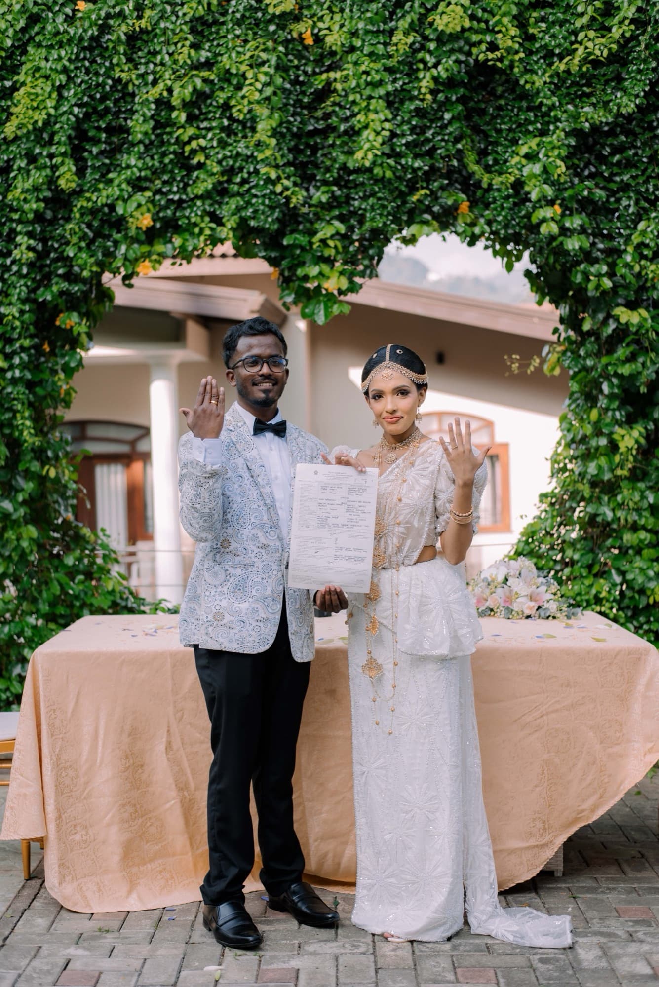 Couple exchanging rings at a flower-strewn table beneath a leafy archway, a tiled villa and misty mountains behind them in soft daylight.