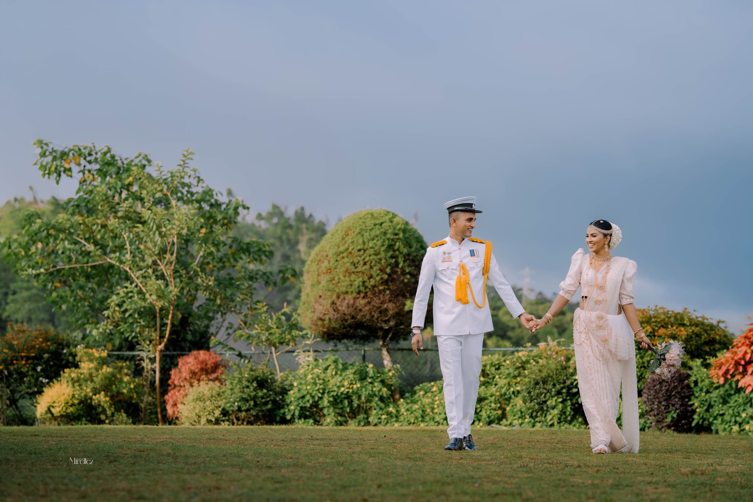A naval-uniformed groom and bride walking hand in hand across a garden at golden hour