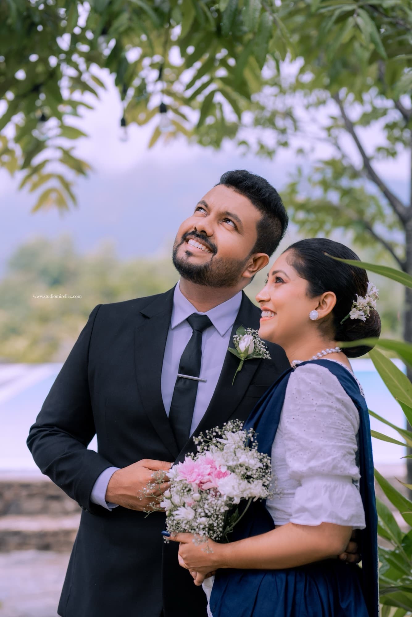 An engaged couple in a black suit and navy saree holding a bouquet outdoors