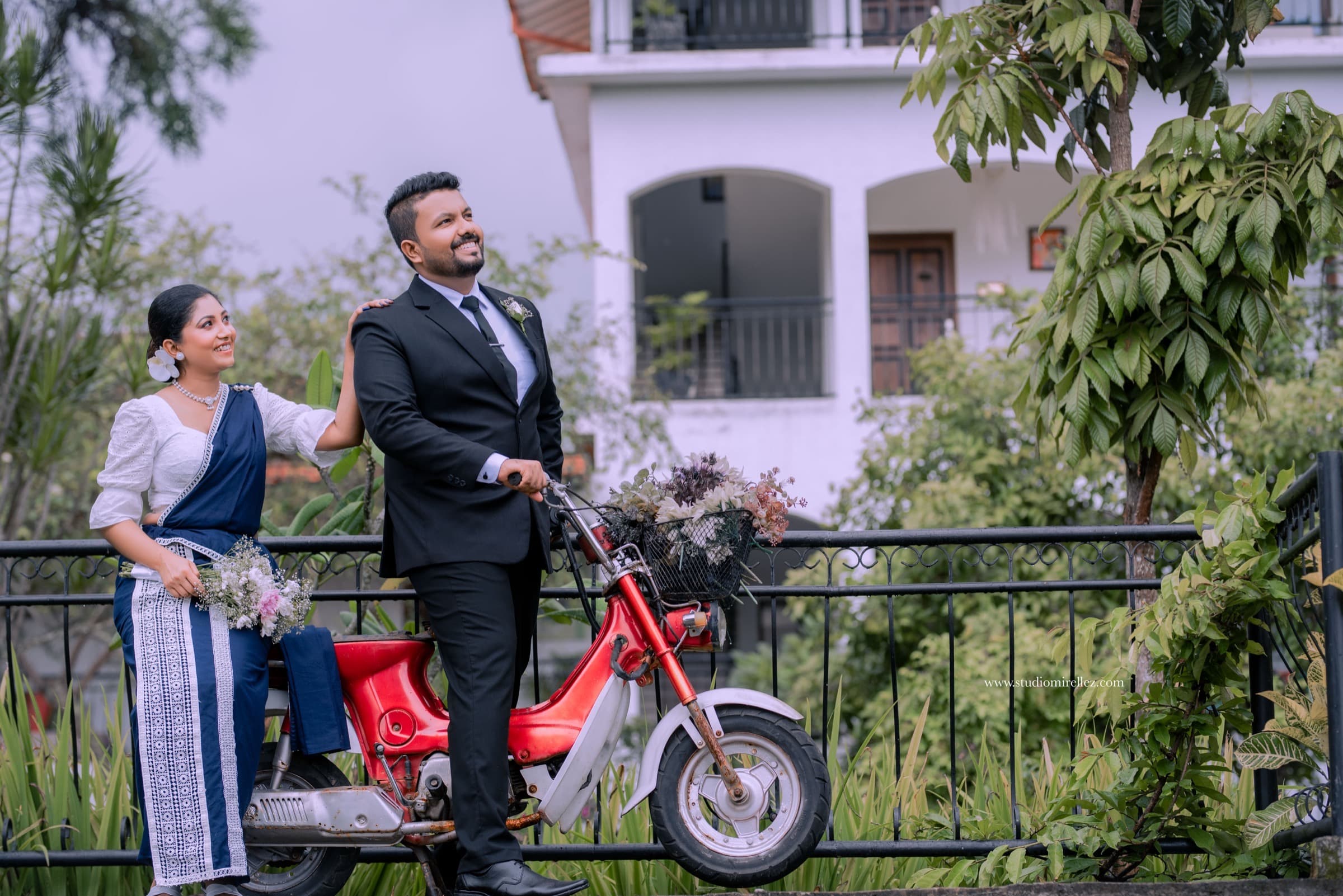 An engaged couple beside a vintage red moped in a lush garden