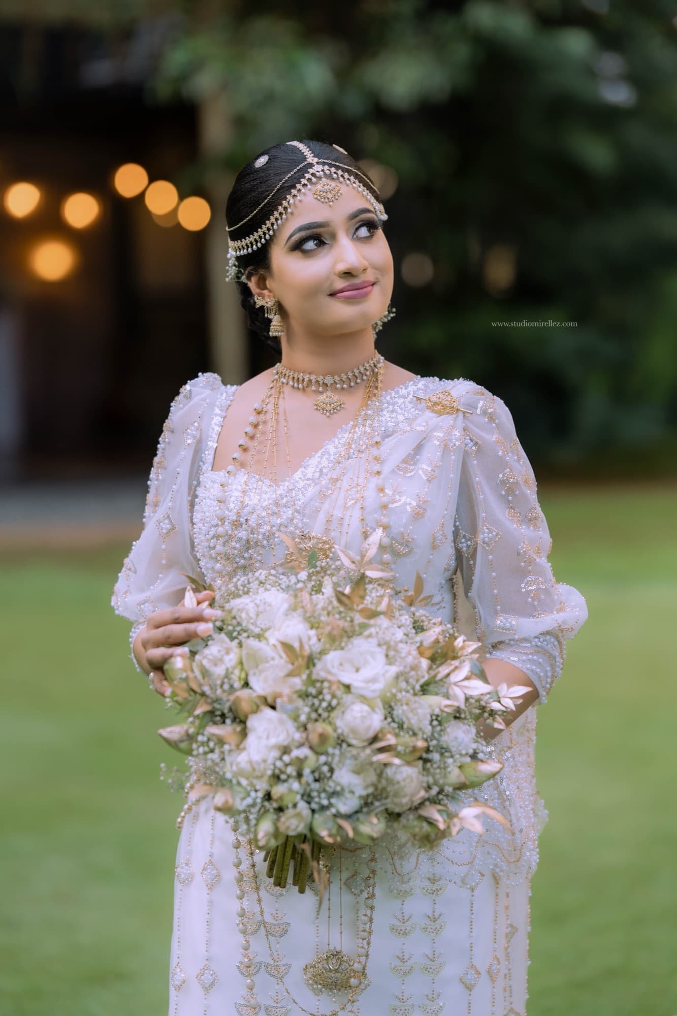 A radiant Kandyan bride in white bridal attire holding a bouquet at golden hour
